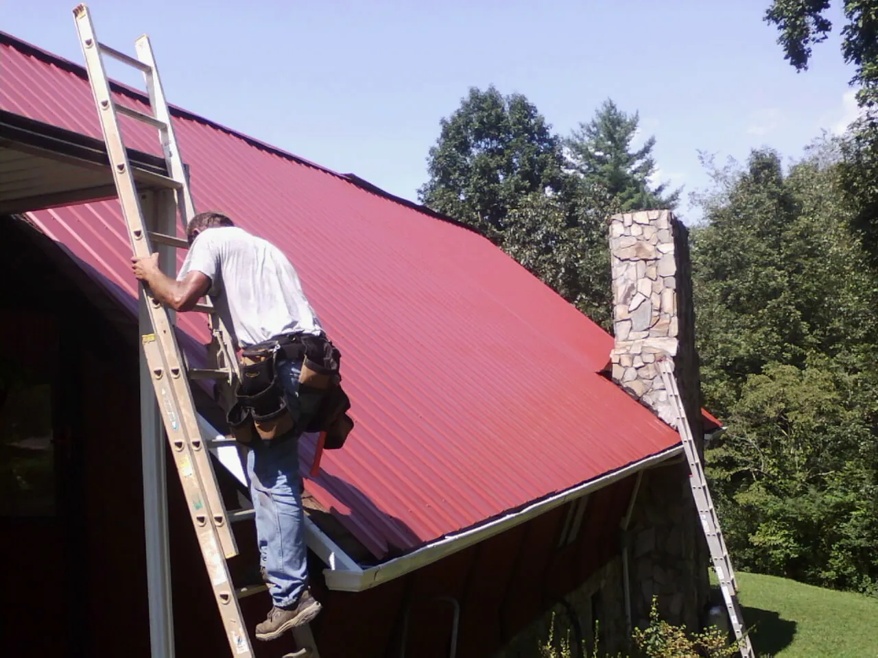 Roofer on a ladder beside a red metal roof and stone chimney — Soffit & Fascia Repair by Roofing Brothers in Middlesex